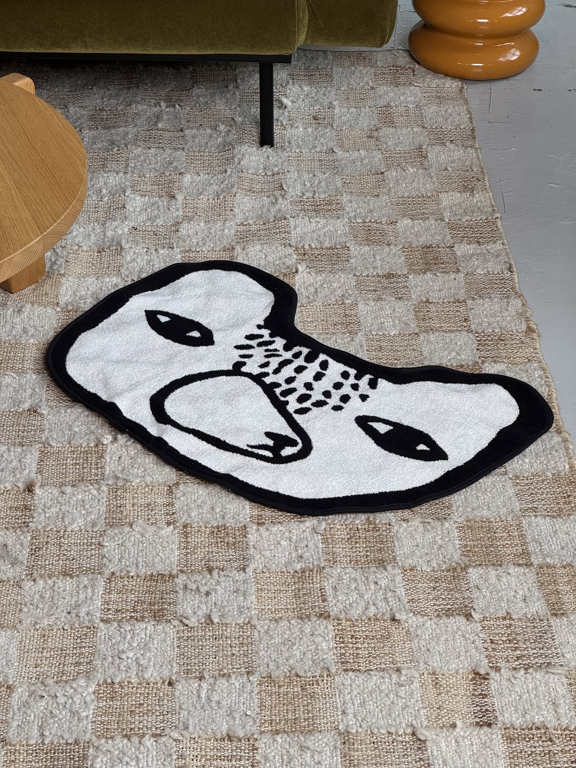 The Donna Wilson Bear Shaped Bath Mat by Donna Wilson, featuring a small white bear design with a black outline and simple face, is placed on a beige checkered carpet near a wooden table and green sofa.