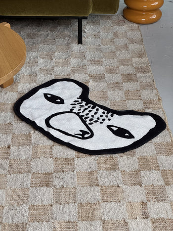 The Donna Wilson Bear Shaped Bath Mat by Donna Wilson, featuring a small white bear design with a black outline and simple face, is placed on a beige checkered carpet near a wooden table and green sofa.