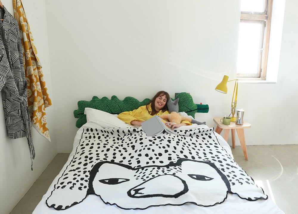 A woman in a yellow shirt reads in bed with a green headboard and the Donna Wilson Bear Shaped Cotton Throw. The bedroom includes a small wooden side table, lamp, and sunlight streaming through the window.