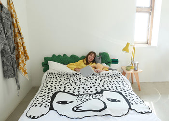 A woman in a yellow shirt reads in bed with a green headboard and the Donna Wilson Bear Shaped Cotton Throw. The bedroom includes a small wooden side table, lamp, and sunlight streaming through the window.