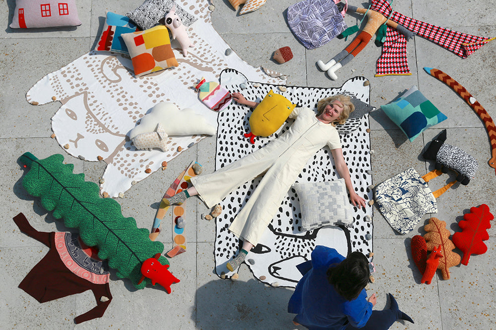 A woman in a cream jumpsuit lies on a Donna Wilson Bear Shaped Cotton Throw, surrounded by whimsical cushions and plush toys, while another person in blue kneels nearby, creating a playful and artistic scene.