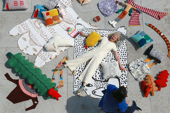 A woman in a cream jumpsuit lies on a Donna Wilson Bear Shaped Cotton Throw, surrounded by whimsical cushions and plush toys, while another person in blue kneels nearby, creating a playful and artistic scene.