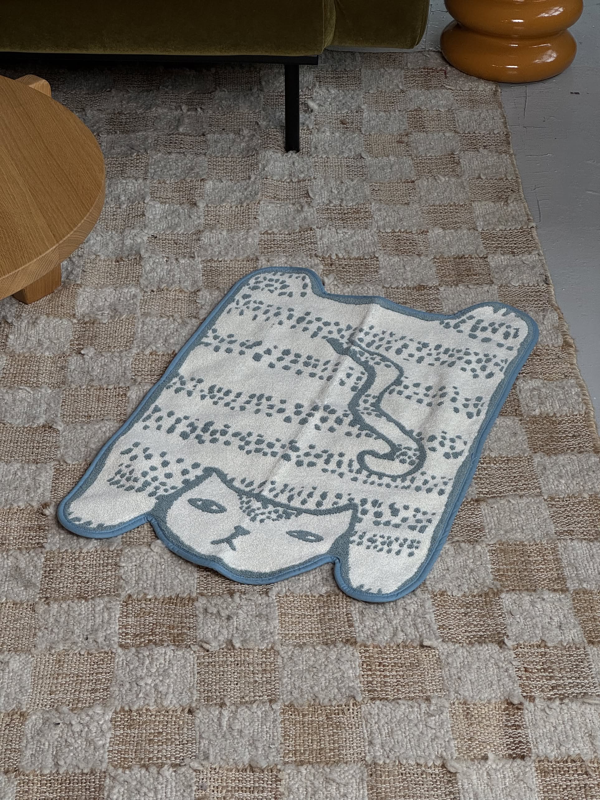 The Donna Wilson Blue Cat Bath Mat by Donna Wilson is a small, rectangular cotton mat with a playful white cat design, curled tail, relaxed expression, and blue border, shown on a larger checkered rug near a wooden table and green sofa.