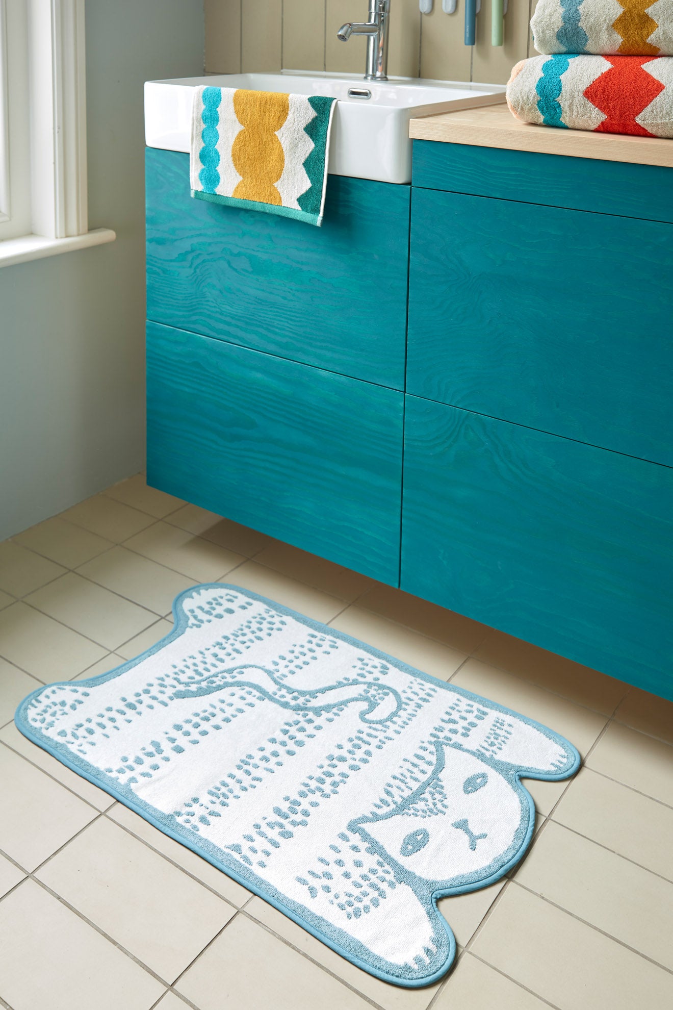 A Donna Wilson Blue Cat Bath Mat by Donna Wilson, made from soft cotton, sits on a beige tiled floor near a teal cabinet, with a matching scallop-patterned towel over the sink—perfect for blue cat-themed bathroom decor.