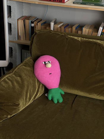 A Donna Wilson Creature - Claude plush from Donna Wilson, in pink and green with a face, leans against an olive-green velvet couch; books are arranged on a shelf in the background.