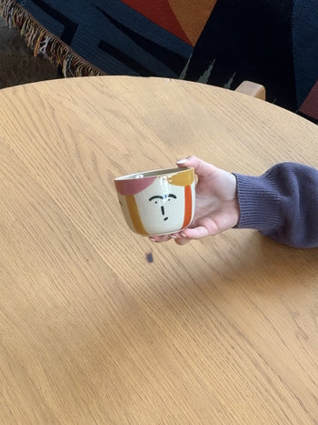 A person with a purple sleeve holds the Donna Wilson Freya Face Cup, a hand-painted stoneware coffee cup by Donna Wilson, over a light wooden table.