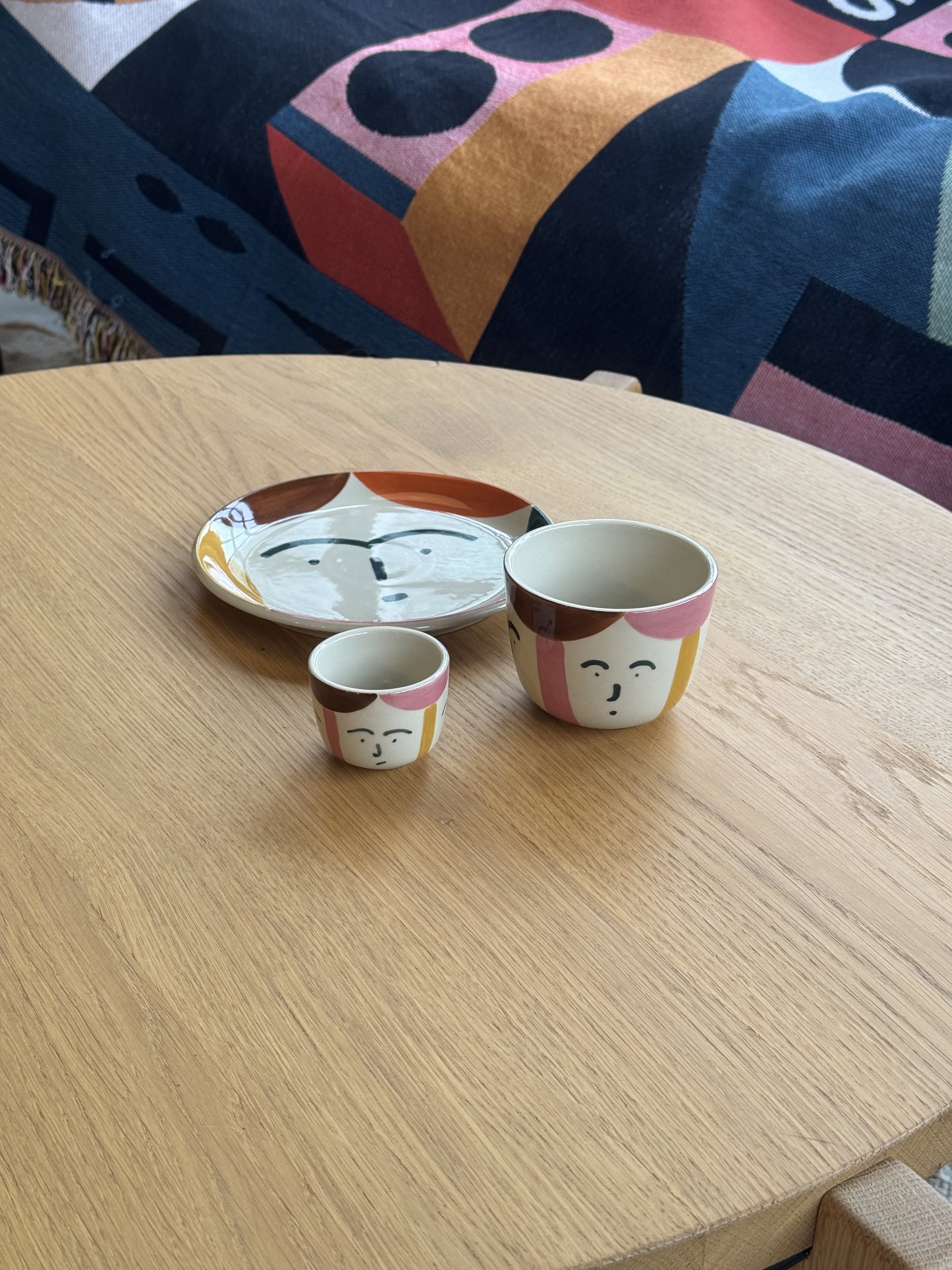 A Donna Wilson Freya Face Cup, a medium artisanal coffee cup, and a plate with abstract face designs sit on a light wooden table, with colorful geometric-patterned fabric in the background.