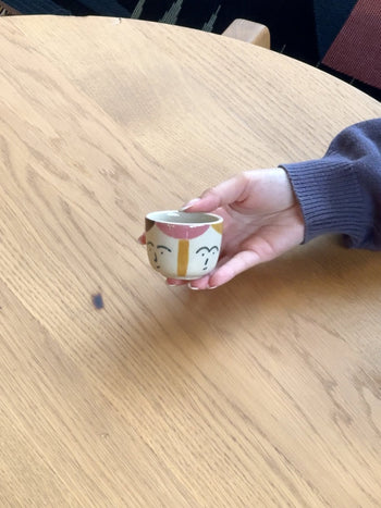 A hand in a purple sleeve holds the Donna Wilson Freya Face Pinch Pot—a small ceramic cup with a simple hand-painted stoneware face—over a light wooden table marked by a subtle grain.