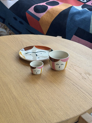 The Donna Wilson Freya Face Pinch Pot, featuring a hand-painted illustrated face, sits with other stoneware pieces on a light wooden table, complemented by Donna Wilson’s colorful abstract-patterned blanket in the background.