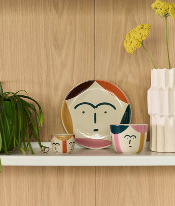 A shelf displays a green plant, a ceramic plate, two Donna Wilson Freya Face Pinch Pots with abstract faces, and a textured white vase holding yellow flowers, all set against a light wood background.