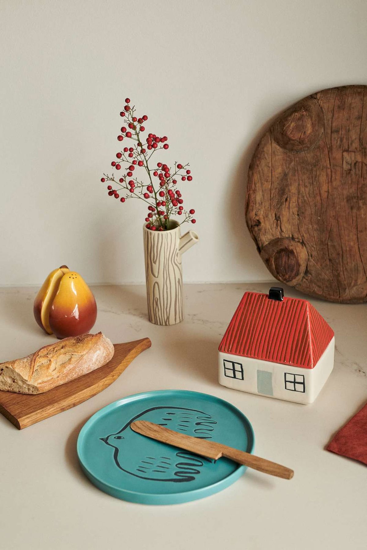 A Donna Wilson Small Log Jug / Vase shaped like a tree branch with red berries sits on a counter next to a house-shaped jar, a teal bird-illustrated plate, and a pear-shaped container.