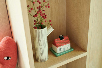 A Donna Wilson Small Log Jug, shaped like a tree trunk with red berries, rests next to a house-shaped ornament on a wooden shelf, adding woodland charm to the display.