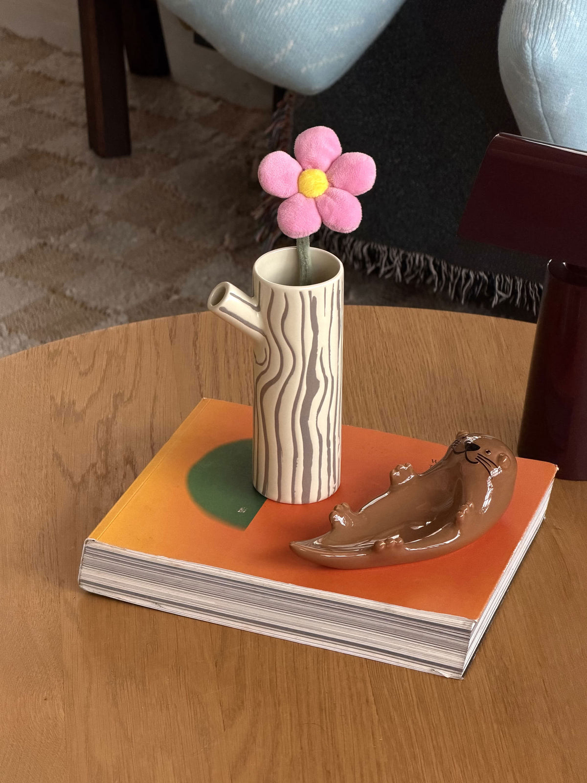 A cylindrical Donna Wilson Small Log Jug with a tree-bark pattern and side spout, holding a pink plush flower, sits on an orange book next to a small brown sea otter figurine.