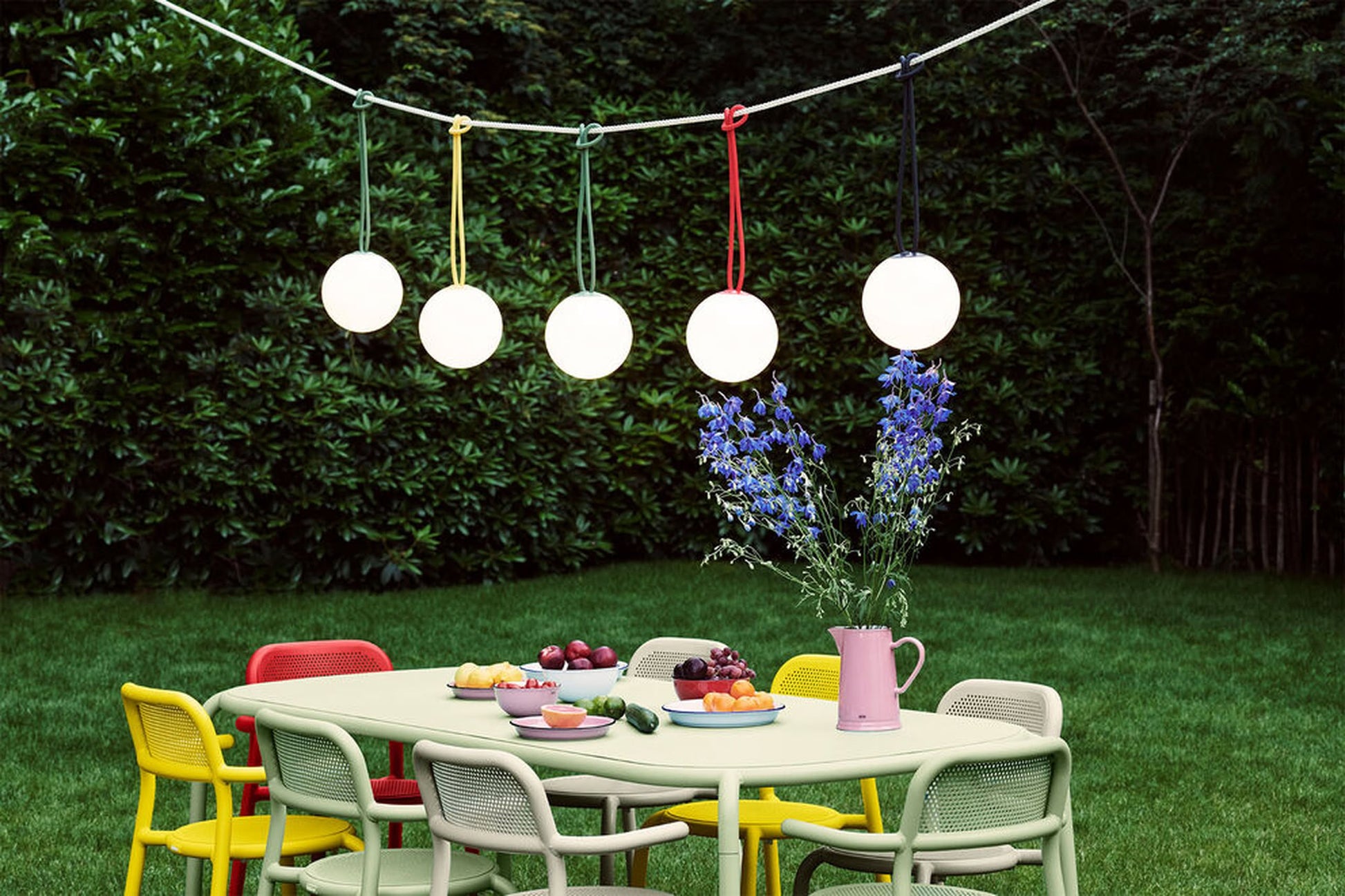 A colorful outdoor dining table with chairs is set on grass, topped with fruit and flowers in a pink vase. Five Fatboy Bolleke lights by Fatboy hang above the table, providing soft glow amid lush greenery.