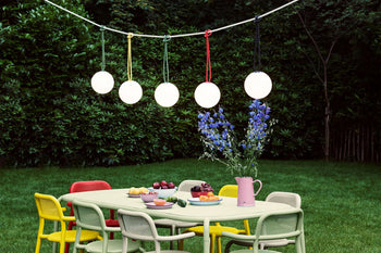 A colorful outdoor dining table with chairs is set on grass, topped with fruit and flowers in a pink vase. Five Fatboy Bolleke lights by Fatboy hang above the table, providing soft glow amid lush greenery.