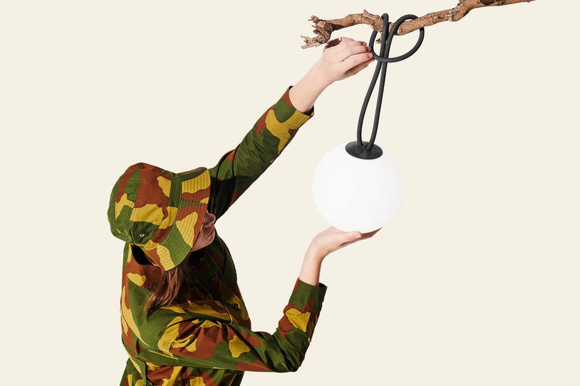 A person in green and brown camo hangs the Fatboy Bolleke, a round white portable light by Fatboy, from a branch against a light background, creating unique outdoor lighting.