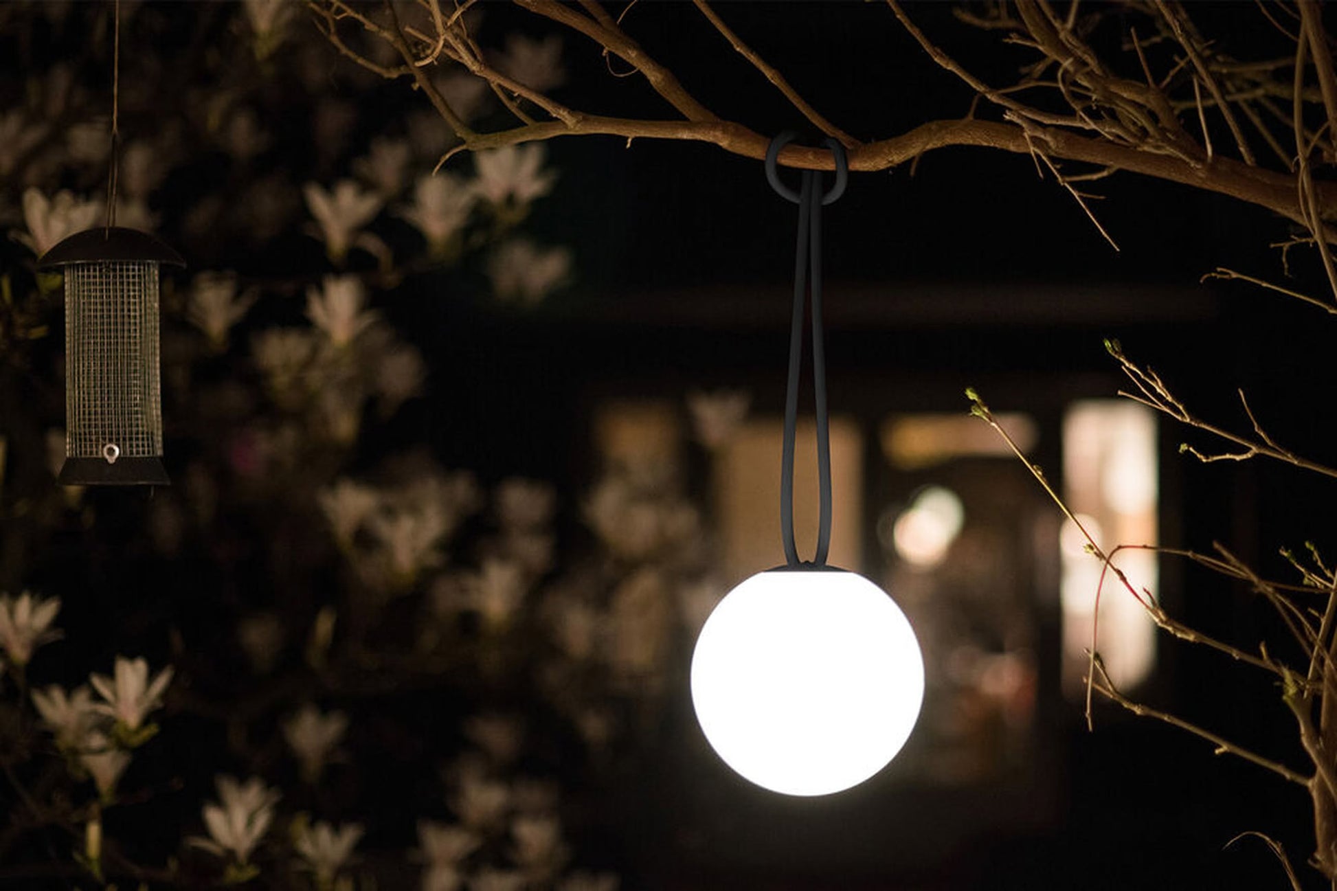 The Fatboy Bolleke by Fatboy, a round glowing portable light, hangs from a tree branch beside a bird feeder at night, casting inviting ambient lighting with soft-focus white flowers and a warmly lit building in the background.