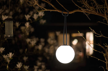 The Fatboy Bolleke by Fatboy, a round glowing portable light, hangs from a tree branch beside a bird feeder at night, casting inviting ambient lighting with soft-focus white flowers and a warmly lit building in the background.