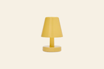 A compact, modern yellow Fatboy Edison The Ambiance LED wireless lamp with a round base and conical shade, set against a plain beige background. Inspired by Fatboy’s playful rechargeable table lamp design.