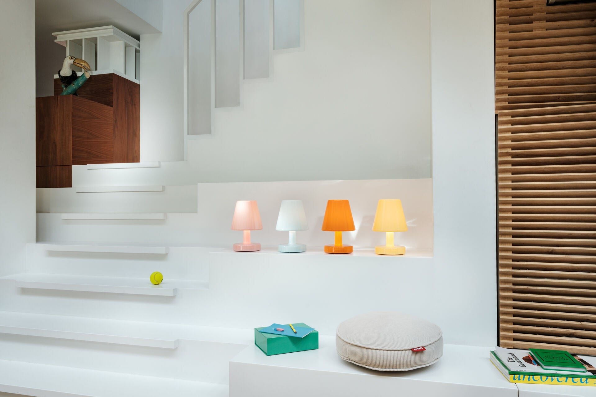 A modern white room with wooden accents features three Fatboy Edison The Ambiance rechargeable lamps on a ledge, a beige round cushion, a green book, stairs, and a wooden bird sculpture on a cabinet.