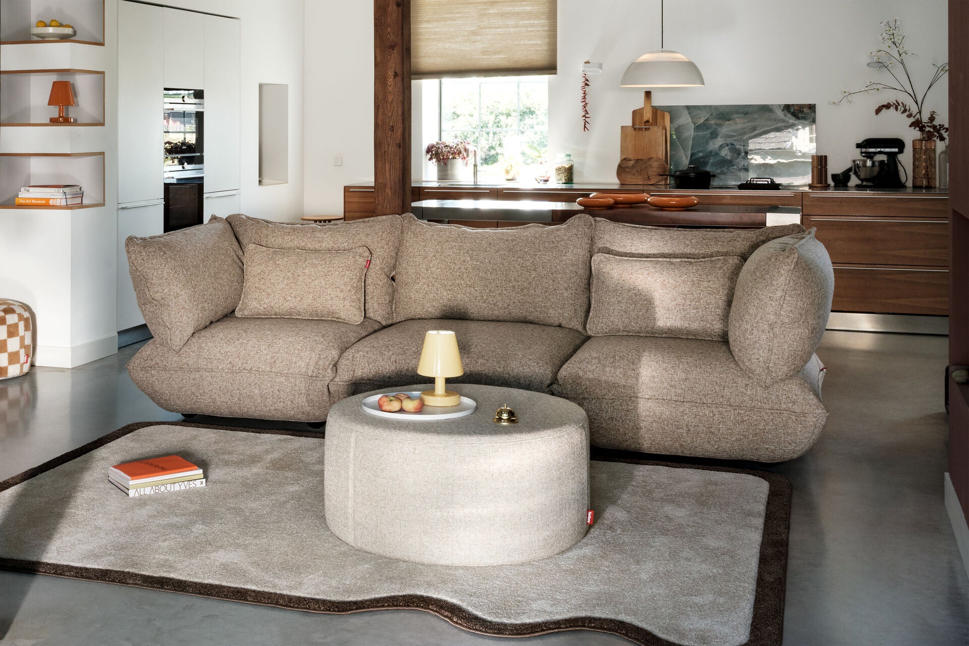 A modern living room with a textured beige sofa, a round ottoman featuring the Fatboy Edison The Ambiance LED lamp and fruit plate, a light rug, and an open kitchen in the background with wooden cabinets and a window.