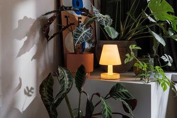 A yellow Fatboy Edison The Ambiance LED wireless lamp by Fatboy glows on a white shelf among potted green plants, casting plant shadows on the wall. In the background, a poster with abstract shapes and text is partly visible.