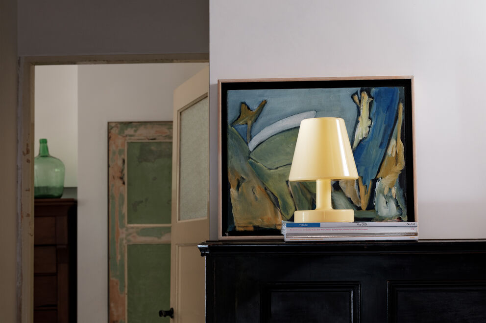 The Fatboy Edison The Ambiance rechargeable table lamp by Fatboy, in yellow, sits atop two books before an abstract painting on a dark wooden cabinet. Open doors behind reveal a green cabinet and a glass bottle.