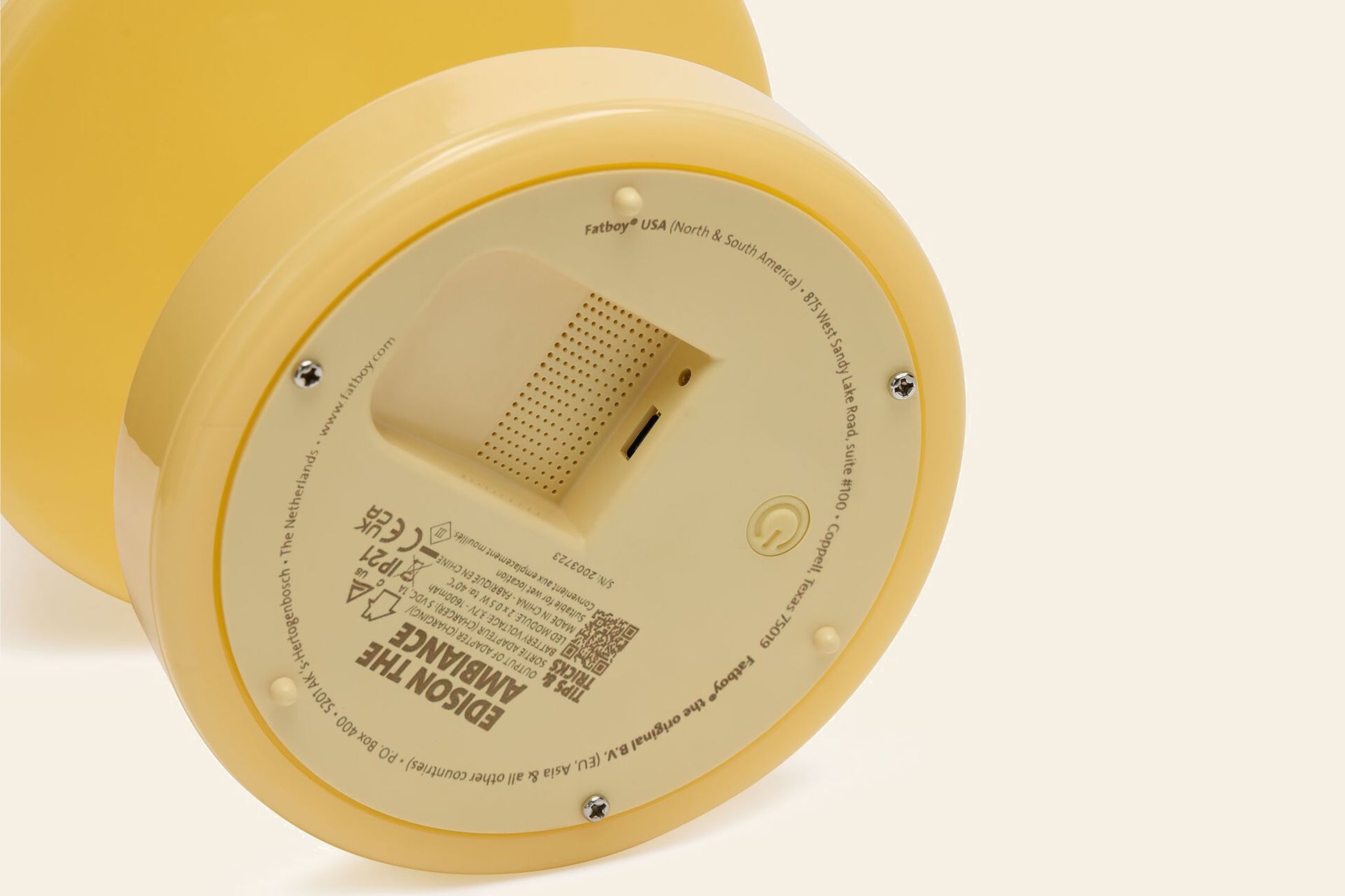 Close-up of the bottom of a yellow, round Fatboy Edison The Ambiance LED wireless lamp showing the power button, speaker grille, product info, certification labels, and battery compartment.