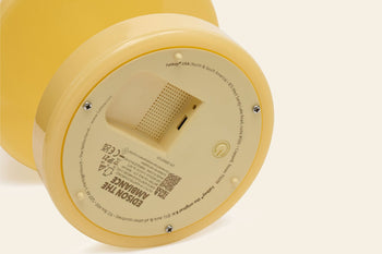 Close-up of the bottom of a yellow, round Fatboy Edison The Ambiance LED wireless lamp showing the power button, speaker grille, product info, certification labels, and battery compartment.