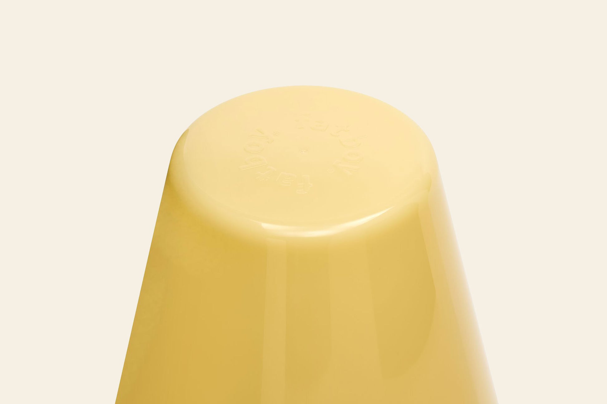 A close-up of an upside-down yellow plastic cup resembling the smooth base of a Fatboy Edison The Ambiance rechargeable table lamp by Fatboy, set against a plain beige background with faint embossed text.