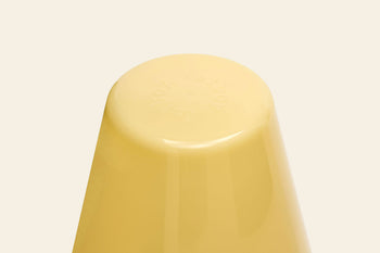 A close-up of an upside-down yellow plastic cup resembling the smooth base of a Fatboy Edison The Ambiance rechargeable table lamp by Fatboy, set against a plain beige background with faint embossed text.