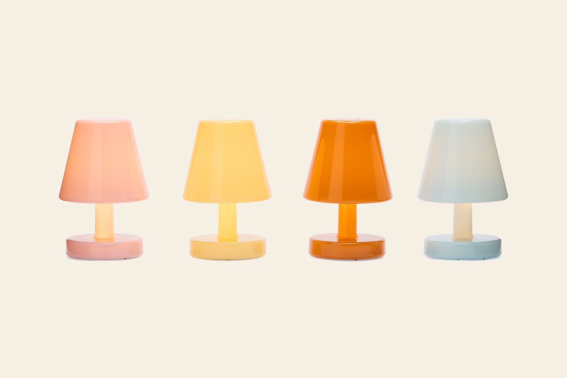 Four Fatboy Edison The Ambiance rechargeable LED table lamps in pastel pink, yellow, orange, and light blue are arranged in a row on a beige background. Each features a simple, modern design with matching base and shade.