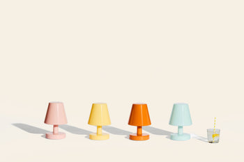 Four Fatboy Edison The Ambiance rechargeable table lamps in pastel pink, yellow, orange, and light blue are lined up on a light surface. A glass of water with a yellow-striped straw stands to the right against a beige background.