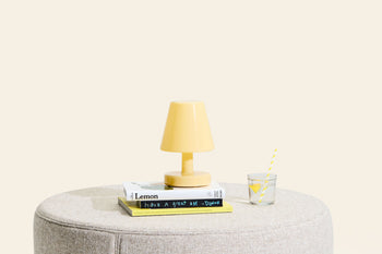 A compact Fatboy Edison The Ambiance wireless lamp by Fatboy, a stack of books, and a glass of water with lemon and straw rest on a round beige ottoman set against a plain light background.