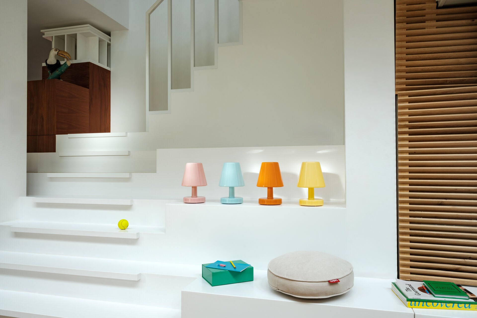 A modern white staircase with four Fatboy Edison The Ambiance wireless LED lamps (blue, pink, orange, yellow) on a ledge, a beige cushion, green box, books nearby; a bird sculpture sits on a wooden cabinet under the steps.