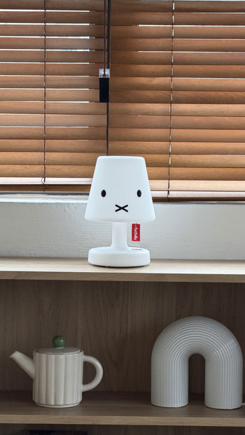 A white Fatboy Edison the Petit x Miffy rechargeable LED lamp sits on a shelf in front of wooden blinds, while a ceramic teapot and arched décor feature below.