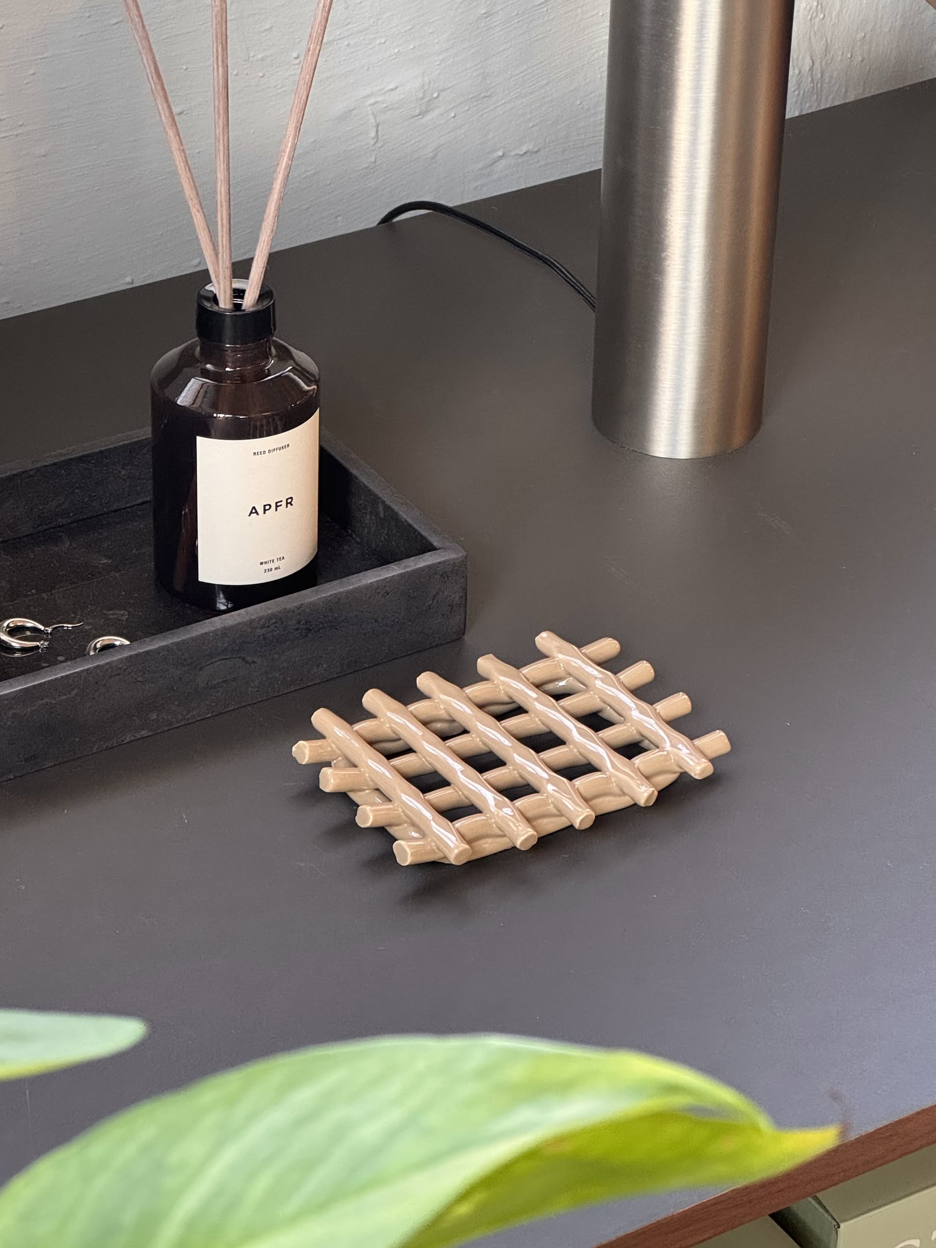 A dark tabletop holds an APFR brown reed diffuser, a black tray with keys, a metallic lamp base, and the Ferm Living Ceramic Soap Tray in Cashmere lattice style. A green plant in the foreground adds freshness.