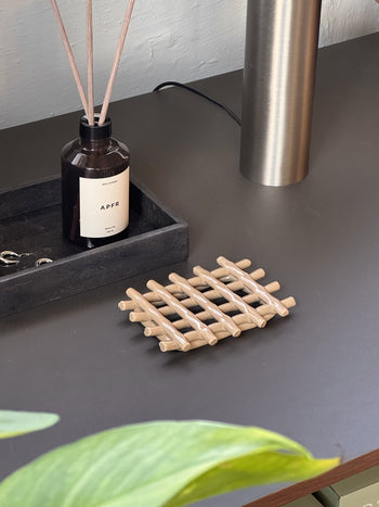 A dark tabletop holds an APFR brown reed diffuser, a black tray with keys, a metallic lamp base, and the Ferm Living Ceramic Soap Tray in Cashmere lattice style. A green plant in the foreground adds freshness.