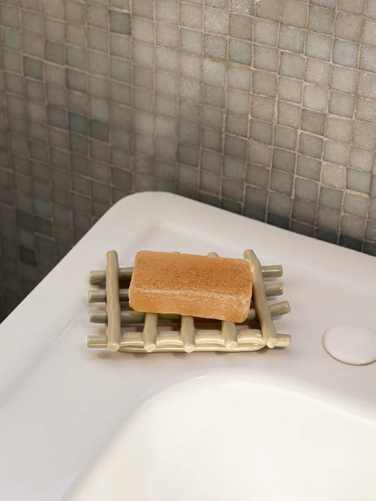 A rectangular bar of brown soap sits on the Ferm Living Ceramic Soap Tray - Cashmere by Ferm Living, positioned on the edge of a white sink with gray mosaic tiles in the background.