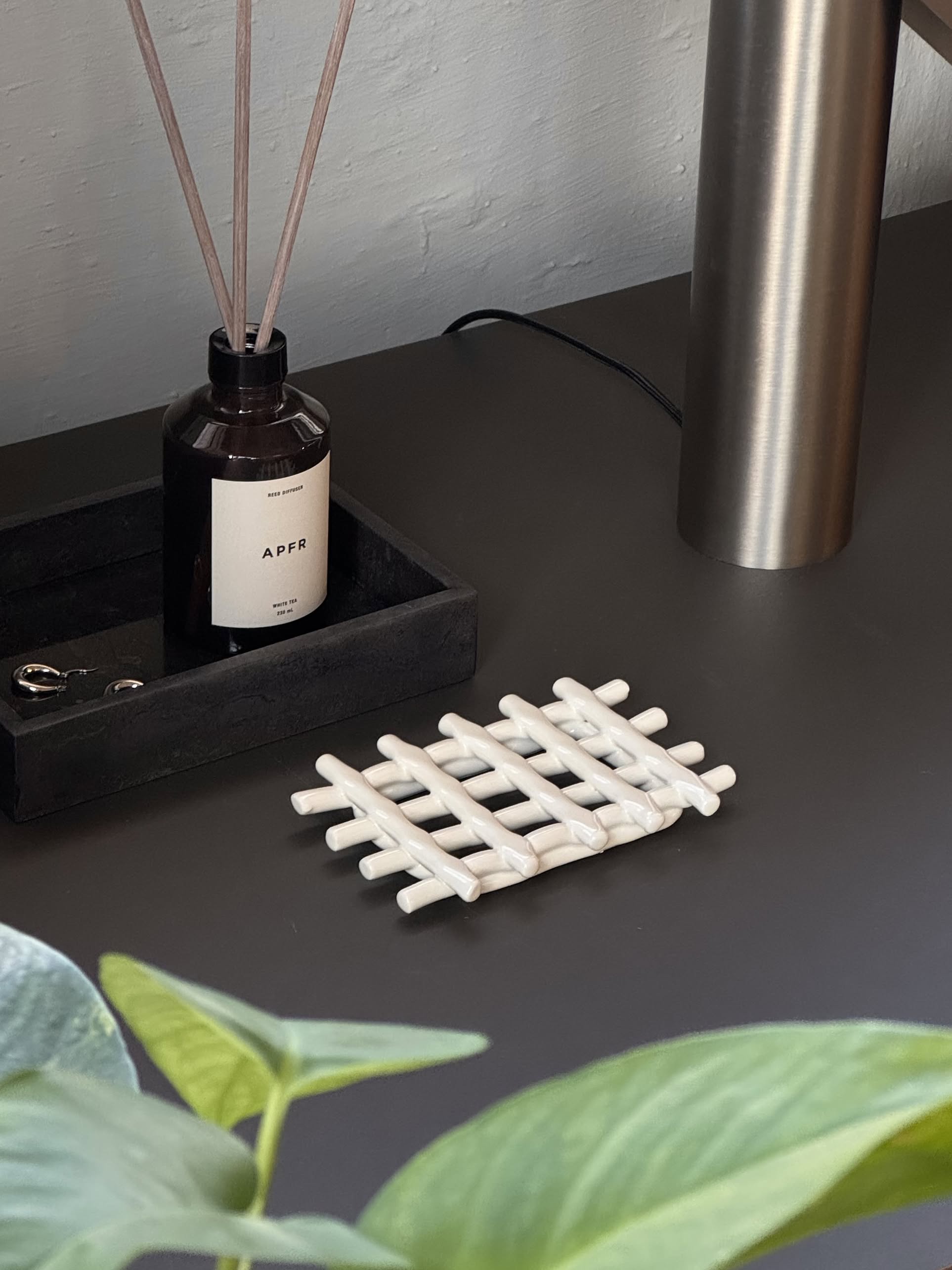 A dark tabletop features a black tray with a brown APFR diffuser and reeds, beside a modern metal lamp base and the Ferm Living Ceramic Soap Tray in off-white. Green leaves are visible in the foreground.