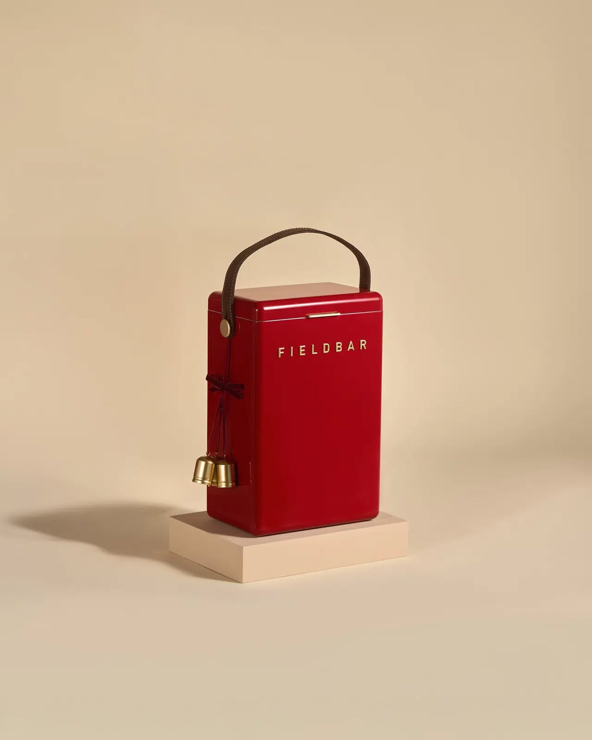 A red Fieldbar Drinks Box by Fieldbar with a dark brown handle and two small gold bells tied with red string rests on a light beige platform against a plain beige background.