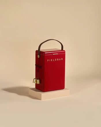 A red Fieldbar Drinks Box by Fieldbar with a dark brown handle and two small gold bells tied with red string rests on a light beige platform against a plain beige background.