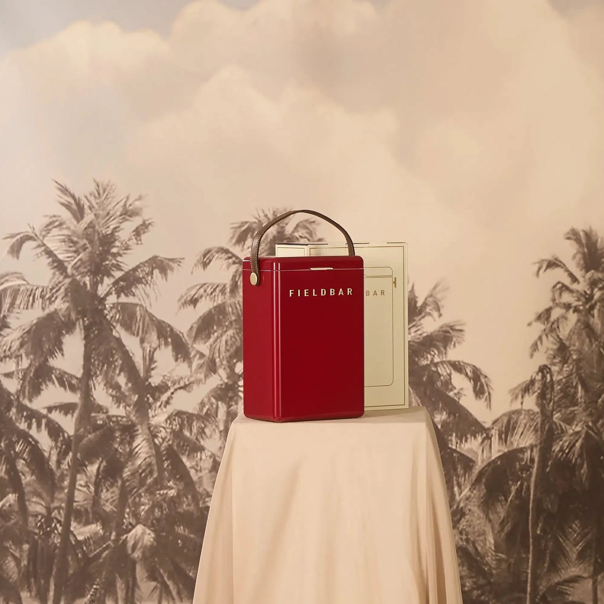 A red Fieldbar Drinks Box by Fieldbar, featuring a handle, rests on a fabric-covered table against a sepia backdrop with palm tree silhouettes and a cloudy sky.