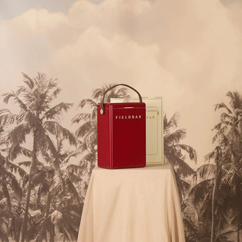 A red Fieldbar Drinks Box by Fieldbar, featuring a handle, rests on a fabric-covered table against a sepia backdrop with palm tree silhouettes and a cloudy sky.