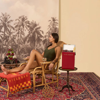A woman in a dark swimsuit relaxes on a wicker lounge chair with a drink, beside a red Fieldbar Drinks Box by Fieldbar on a small table, surrounded by tropical decor, a patterned rug, and palm trees.