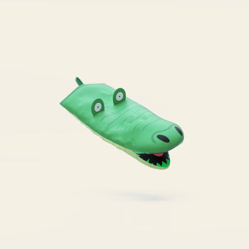 The Fluid Market Crocodile Oven Mitt, a playful green kitchen accessory with cartoon eyes and an open mouth, floats against a plain light background. This whimsical puppet-like mitt is part of the Fluid Market x Tiffany Cooper collaboration.