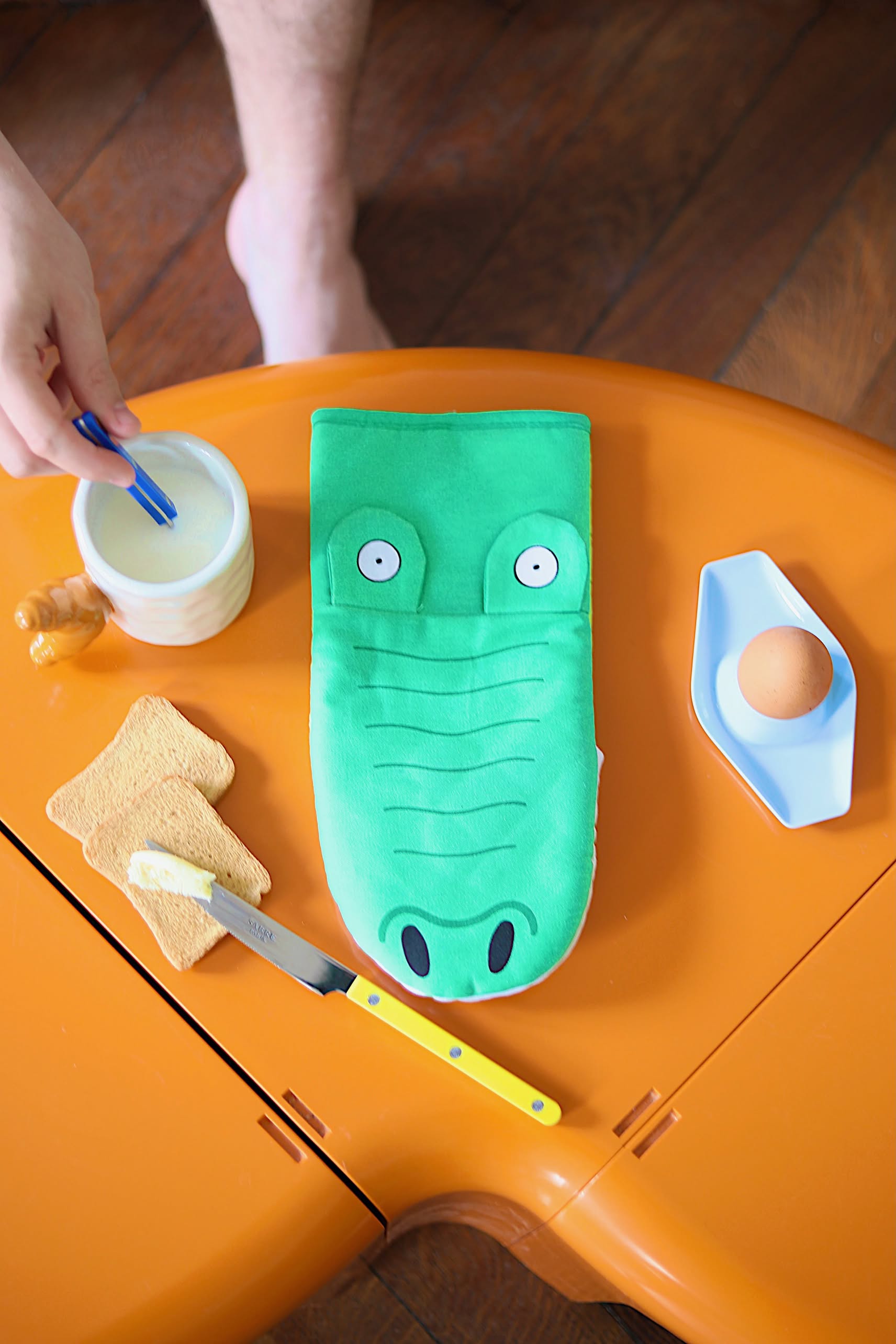 A person dips a spoon into a cup near an orange table with the Fluid Market Crocodile Oven Mitt, two pieces of toast, a knife, an egg in a blue dish, and a honey stick.
