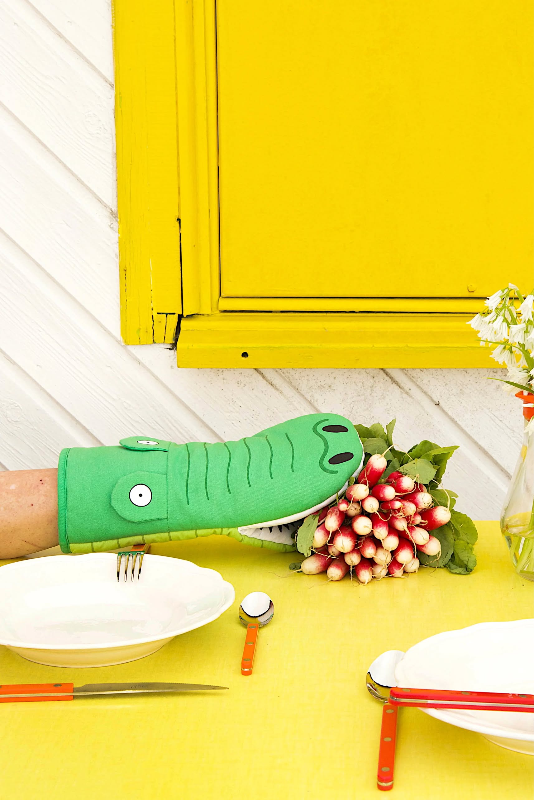 A person’s arm wearing the Fluid Market Crocodile Oven Mitt reaches for radishes on a yellow table set with plates and cutlery, against a white wall with a bright yellow window and flowers in a jar.