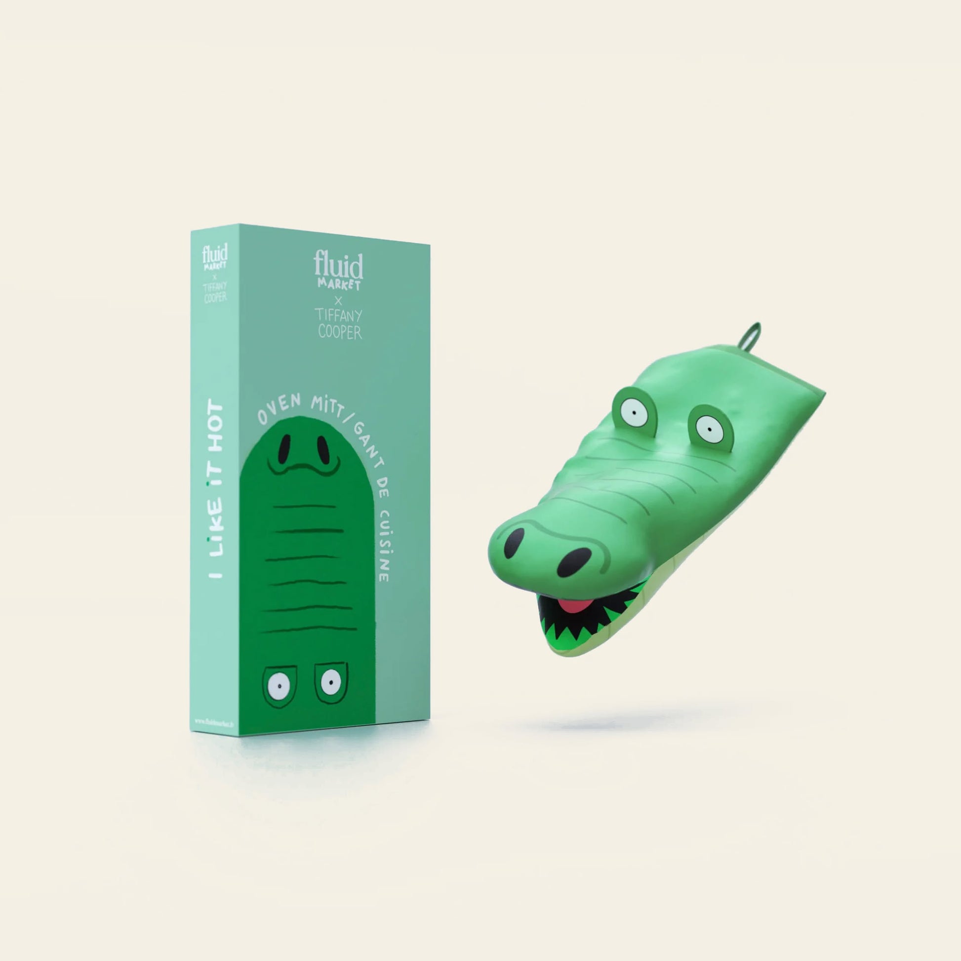 The Fluid Market Crocodile Oven Mitt is shown beside its matching teal box featuring a crocodile illustration and the text “I LIKE IT HOT”—a playful kitchen accessory from Fluid Market.
