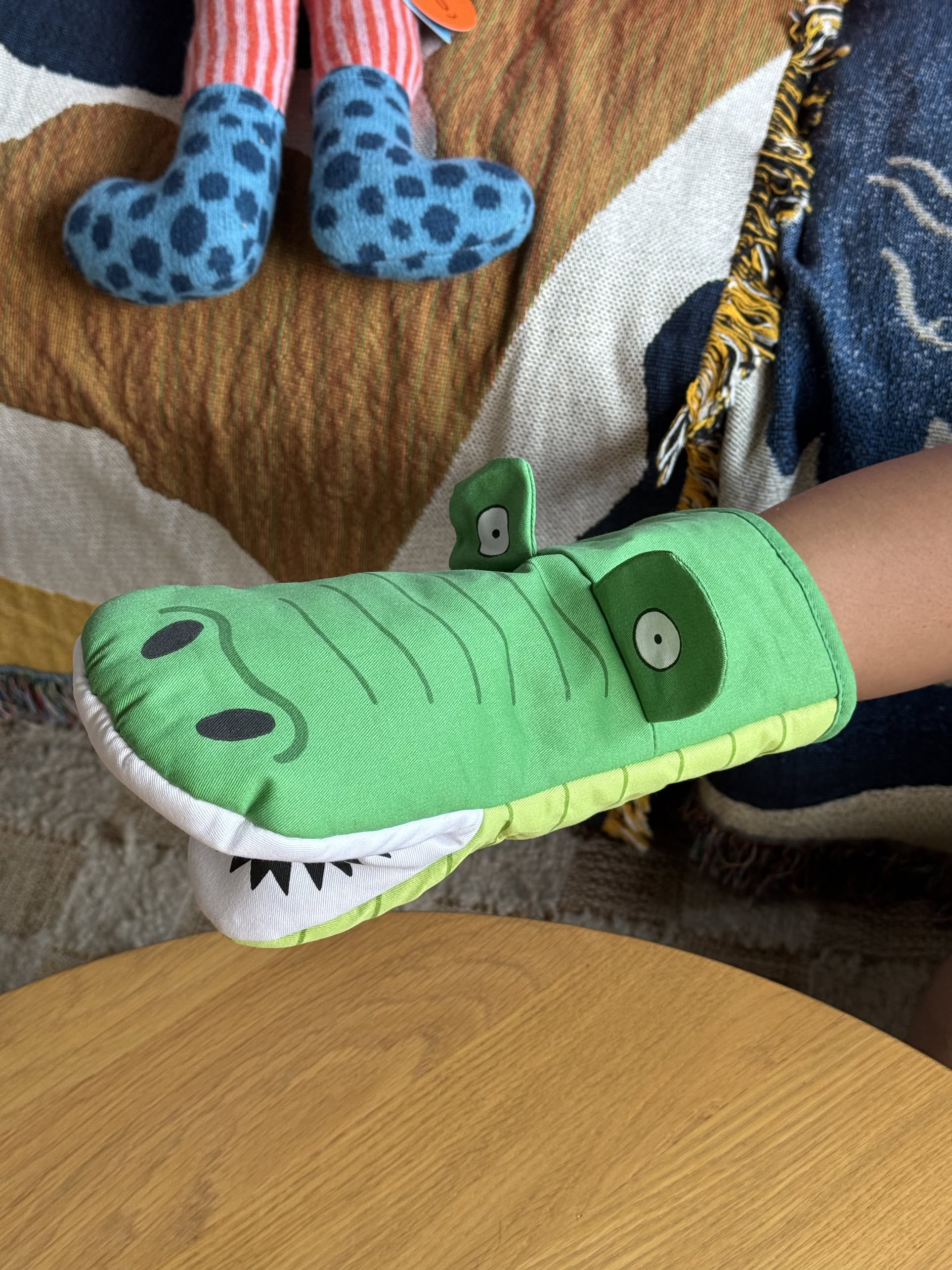 A person wears the Fluid Market Crocodile Oven Mitt by Fluid Market on their hand above a light wooden surface, with a blue spotted stuffed animal and a brown patterned blanket in the background.